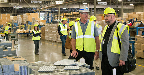 People walking through a plant in high visibility vests and hard hats and two men standing next to truss plates